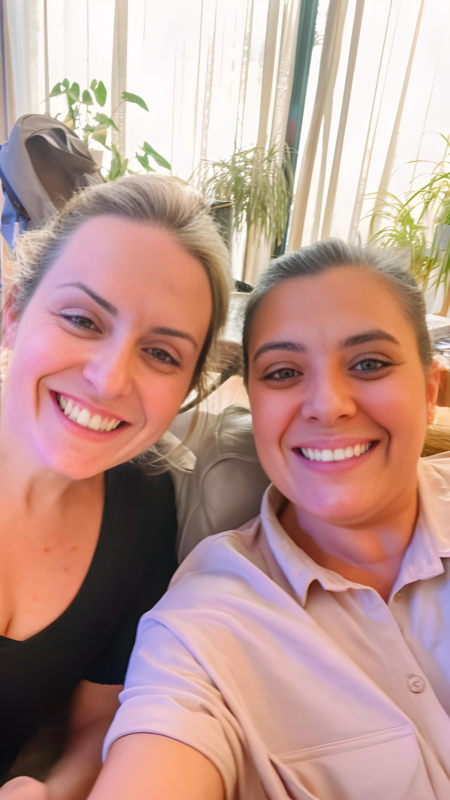 Two smiling women taking a close-up selfie indoors. They are seated on a couch, with natural light coming through large windows and several green potted plants in the background. Both are casually dressed and appear happy and relaxed.
