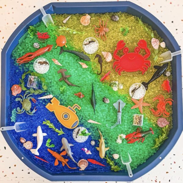 Colorful ocean-themed sensory tray filled with blue-to-green-to-yellow beads, plastic sea creatures (crabs, starfish, sharks, squid), shells, and scooping cups for kids to play.