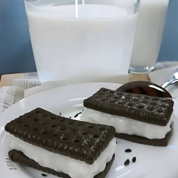 Two chocolate sandwich cookies with white cream filling on a white plate, with a glass of milk in the background.