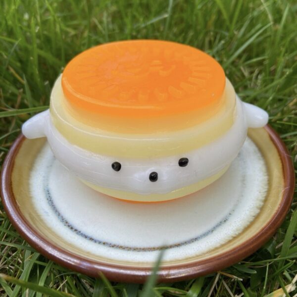 Whimsical orange-and-white toy cake stacked on a brown ceramic plate in grass, with a small face of black eyes on the white middle ring.